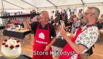 Billedet viser en livlig scene fra "Den Store Kagedyst" under et teambuilding-arrangement i Aarhus. To mænd, klædt i røde forklæder med BoligViborg-logoet, ser ud til at have det sjovt og dele et øjeblik af glæde og triumf med publikummet. Den ene mand løfter en kage op, mens den anden klapper og griner. Publikummet, bestående af en blanding af ældre og yngre voksne, ser ud til at nyde forestillingen. Denne type aktivitet er perfekt til at styrke samarbejde og skabe en afslappet atmosfære, hvor alle kan deltage og have det sjovt.