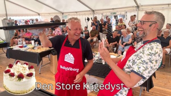 Billedet viser en livlig scene fra "Den Store Kagedyst" under et teambuilding-arrangement i Aarhus. To mænd, klædt i røde forklæder med BoligViborg-logoet, ser ud til at have det sjovt og dele et øjeblik af glæde og triumf med publikummet. Den ene mand løfter en kage op, mens den anden klapper og griner. Publikummet, bestående af en blanding af ældre og yngre voksne, ser ud til at nyde forestillingen. Denne type aktivitet er perfekt til at styrke samarbejde og skabe en afslappet atmosfære, hvor alle kan deltage og have det sjovt.