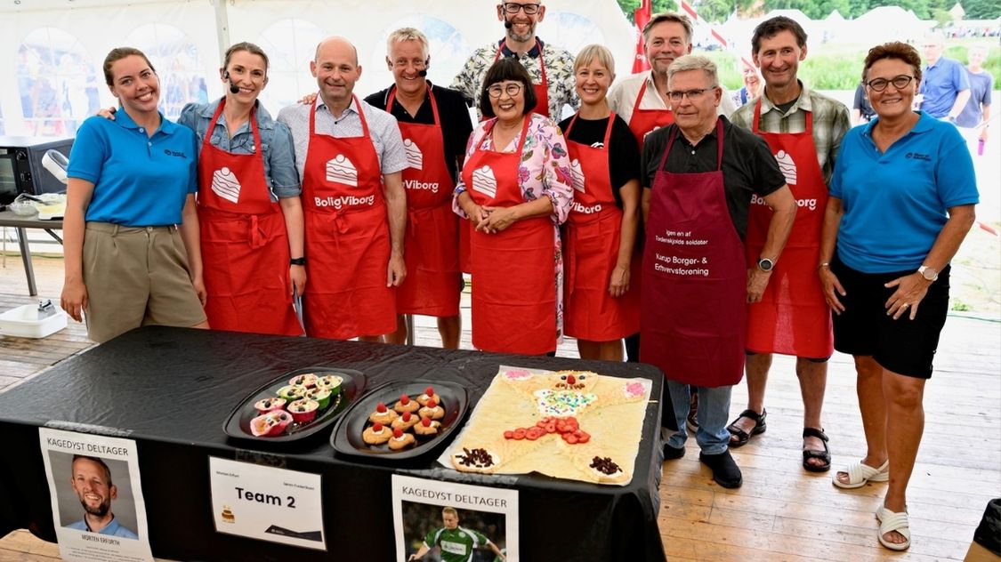 På billedet ser vi et teambuilding-event i Aarhus, hvor deltagere har samlet sig til en "Den Store Kagedyst" aktivitet. Gruppen består af mænd og kvinder i alle aldre, klædt i farverige forklæder, der repræsenterer forskellige lokale organisationer som "BoligViborg". De står stolt bag deres kulinariske kreationer, som er præsenteret på et bord foran dem. Denne type aktivitet fremmer samarbejde og kreativitet, idet deltagerne arbejder sammen i teams for at skabe de mest indbydende og velsmagende desserter. Det er en sjov og engagerende måde at styrke relationer og teamånd på.