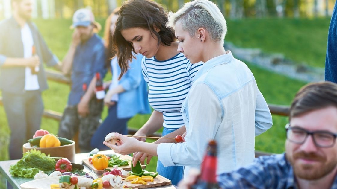 Billedet viser en Outdoor Cooking Challenge, hvor deltagerne arbejder sammen om at forberede og tilberede mad i det fri. To kvinder skærer grøntsager og sætter dem på grillspyd, mens andre deltagere hygger sig i baggrunden. Stemningen er afslappet og social, hvilket gør aktiviteten ideel til teambuilding.